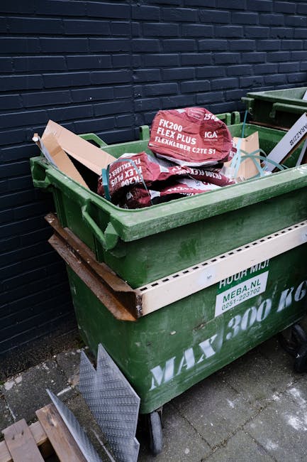 A pile of mixed waste is accumulated on a gravel surface adjacent to a stone wall and a metal pole. The waste includes black plastic rubbish bags, partially opened with visible contents, and an old, d