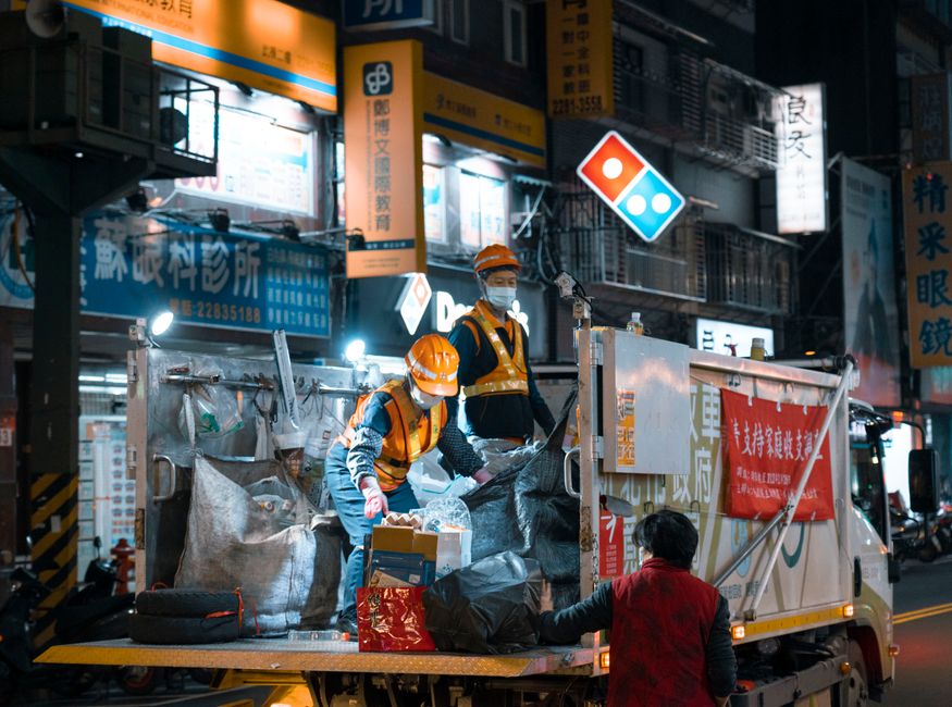 Two waste collection workers wearing orange helmets, reflective vests, and face masks are standing on the open back of a white rubbish collection truck parked on an urban street at night. The truck's